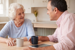 Older woman and man smiling and talking over coffee at a kitchen table, with a tablet resting nearby.